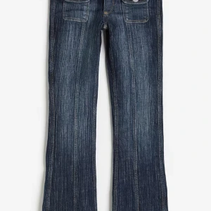 Mörkblå bootcut jeans med fickor - Snygga mörkblå jeans med bootcut passform och markerade vertikala sömmar. Framtill finns två fickor med lock och knappdetaljer. Jeansen har låg midja och är tillverkade i klassiskt denimtyg. Perfekta för dig som gillar retrovibbar och coola detaljer.