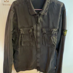 Svart overshirt från Stone Island - Säljer en svart overshirt från Stone Island med klassisk patch på vänster ärm. Jackan har två stora bröstfickor med lock och dragkedja framtill. Tillverkad i bomull och har en clean, stilren look som passar perfekt till streetwear.