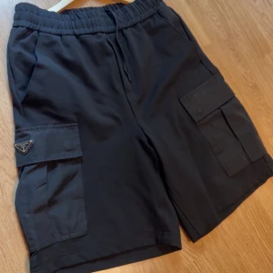 Svarta cargo shorts från Prada - Svarta cargo shorts från Prada med bred resår och snörning i midjan. Stora sidofickor med lock och ikonisk Prada-logga på vänster ficka. Materialet är mjukt och känns som bomull, med detaljer i polyester på fickorna. Perfekt för chill och streetwear.