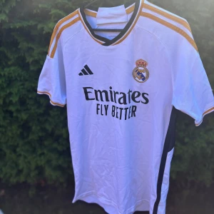Real Madrid hemmatröja Adidas 23/24 - Säljer en officiell Real Madrid hemmatröja från Adidas säsongen 23/24. Tröjan är vit med guld- och marinblå detaljer, broderat klubbmärke och svarta Adidas-loggor. Tillverkad i lätt och ventilerande AEROREADY-material. Klassisk passform med korta ärmar och rund hals. Har ni några frågor är det bara att höra av sig! Pris kan diskuteras👍