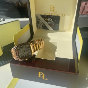 Guldig fyrkantig klocka från Emporio Limited - Snygg klocka från Emporio Limited med fyrkantig urtavla, romerska siffror och guldig länk i metall. Klockan har detaljerade skruvar på armbandet och en vit urtavla med svarta visare. Kommer i originalask med logga och certifikat.  Firenze 2073c    