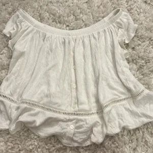 Vit offshoulder blus  - Superfin vit offshoulder blus från Divided i storlek XS. Blusen har knappar framtill, volangdetaljer och en luftig, rynkad design. Perfekt för varma dagar med sin lätta bomullskänsla och snygga, avslappnade passform.