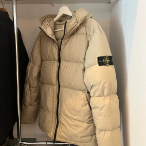 Beige pufferjacka från Stone Island - Snygg beige pufferjacka från Stone Island med ikonisk patch på ärmen. Kvitto och andra bevis på produktens äkthet tillgängliga. Det finns ett litet snitt på jackan vid fickan, men det finns ett annat materiallager bakom det, så till exempel dun kan inte fly genom snittet. Det är också väldigt diskret! 