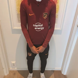 Atletico Madrid Nike Dri-FIT träningströja - Säljer en mörkröd långärmad Nike Dri-FIT träningströja med Atletico Madrid-logga och orange Nike Swoosh. Tröjan har half zip, tight passform och tryckt sponsor på bröstet. Perfekt för fotbollsträning eller sportiga dagar.