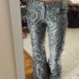 paisleymönstrade byxor - Säljer ett par snygga byxor med svart och silvriga paisleymönster. Byxorna har bootcut-modell med utsvängda ben. Passar någon som har M/L. Midjemått: 45x2                                                             Innerbenslängd: 82