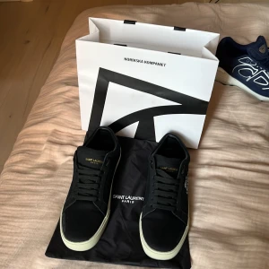 Svarta Saint Laurent sneakers - Svarta sneakers från Saint Laurent med vit kontrasterande sula och handskriven logga på sidan. Skorna har snörning, rund tå och detaljer i skinn och textil. Snygg minimalistisk design med guldfärgad Saint Laurent Paris-tryck på plösen och innersulan.