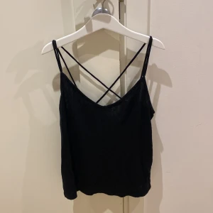 Svart linnetopp från H&M Divided - Svart linnetopp med tunna axelband som korsas i ryggen. Enkel och stilren design, perfekt att matcha med jeans eller kjol. Mjuk och lätt i materialet, med en något lös passform. Snyggt fall och diskret v-ringning framtill.