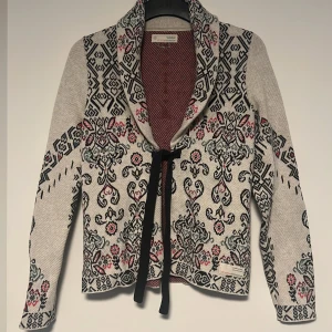 Mönstrad kofta från Odd Molly med knyt - Säljer en snygg kofta från Odd Molly i beige med svart, rött och grått mönster. Koftan har lång ärm, öppen front och ett svart band att knyta med framtill. Materialet är stickat och känns mjukt. Perfekt för dig som gillar detaljer och unika plagg. Använd ca 2 gånger, tvättat 1 gång, mest hängt i garderoben🩷