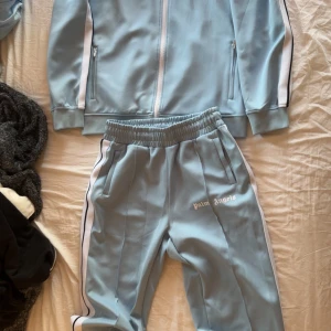 Ljusblå tracksuit från Palm Angels - Säljer en ljusblå track jacket från Palm Angels med vita och svarta ränder längs ärmarna. Jackan har dragkedja framtill, ståkrage och två fickor med dragkedja. Materialet är mjukt och sportigt, perfekt för en chill och trendig look. Tröjan storlek M och byxorna M men passar L bra också