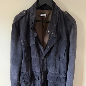 Brunello Cucinelli sude jacka  - Snygg mörkblå mockajacka från Brunello Cucinelli med flera stora fickor framtill, axelklaffar och cool ståkrage med tryckknappar och dragkedja. Fodrad med brunt innerfoder och har en klassisk, passform. Perfekt för dig som gillar stilren och lyxig vibe.