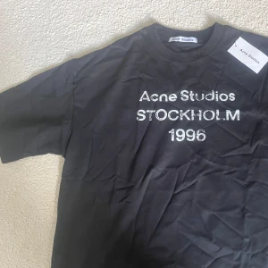 Svart Acne Studios t-shirt Stockholm M - Svart t-shirt från Acne Studios med vit text 'Acne Studios STOCKHOLM 1996' på bröstet. Klassisk rund hals och korta ärmar. Materialet är mjuk bomull och passformen är loose, perfekt för en chill och stilren look. Kommer med tag.