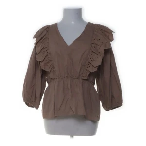 Brun blus med volang från Pernilla Wahlgren - Supersöt brun blus från Pernilla Wahlgren med v-ringning och breda volanger med broderade detaljer över axlarna. Blusen har trekvartsärmar och resår i midjan för en snygg passform. Super fin nu till hösten! Köpt via Sellpy, lappen kvar!