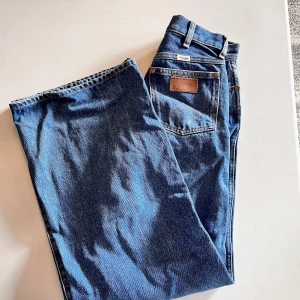 Wrangler wide jeans blå W24 - Säljer ett par klassiska blå jeans från Wrangler med raka, vida ben och hög midja. Jeansen har fem fickor, läderpatch bak och är tillverkade i robust bomullsdenim. Perfekta för dig som gillar en avslappnad och tidlös look.
