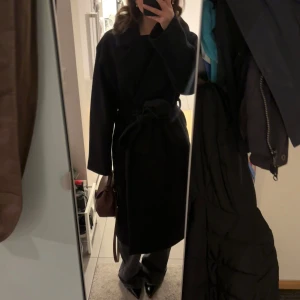 Svart kappa med bälte Kappahl XS - Säljer en svart kappa med bred krage och knytbälte i midjan. Jackan har en rak och oversized passform med långa ärmar, perfekt för dig som gillar en stilren och klassisk look. Materialet känns mjukt och värmande, perfekt för kyligare dagar.