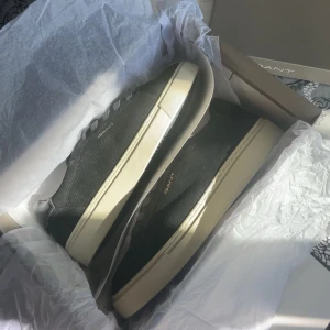 Svarta sneakers från GANT - Snygga svarta sneakers från GANT med klassisk låg profil och rund tå. Skorna har en beige platt sula och är tillverkade i mocka med snörning framtill. GANT-loggan syns i guld på sidan. Perfekta för dig som gillar stilrena och tidlösa sneakers. Nästan aldrig använda, pris kan diskuteras 