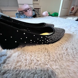 Svarta ballerinaskor med strass från Alley - Snygga svarta ballerinaskor från Alley med spetsig tå och låg klack. Skorna är täckta med glittriga strassdetaljer som ger en extra touch. Utsidan är i mjuk mocka och insidan har en bekväm sula. Perfekta för dig som gillar att sticka ut med stil.