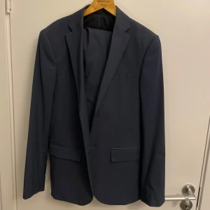 Mörkblå kostym H&M Slim Fit strl 50 - Snygg mörkblå kostym från H&M i slim fit-modell. Kavajen har klassisk design med två fickor med lock och smal krage. Byxorna har raka ben, bälteshällor och bakfickor. Perfekt för dig som gillar stilren och modern look.