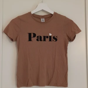 Brun t-shirt Paris print Lindex - Snygg brun t-shirt från Lindex, svart Paris-tryck och vit hjärtdetalj. Rund hals och korta ärmar, tillverkad i mjuk bomull. Storlek 134/140, men kan passa större eftersom den är stretchig!💕 perfekt att växa i. Finns ett minimalt hål på baksidan (se bild) 