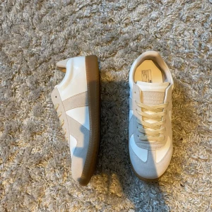 Maison Margiela vita sneakers  - Maison Margiela sneakers i vitt skinn med beige mockadetaljer och klassisk gummisula. Snygg retrostil med rund tå och snörning. Perfekta för dig som gillar minimalistisk och clean look. Märkeslogga under sulan och på plösen.