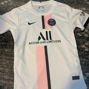 PSG vit och rosa Nike fotbollströja - Snygg PSG fotbollströja från Nike i vitt med en rosa och svart vertikal rand på framsidan. Tröjan har klubbmärke på bröstet, svart Nike-logga och sponsortryck. Tillverkad i lätt och ventilerande material, perfekt för träning eller match.