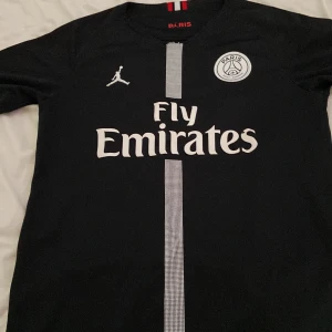 PSG Mbappé svart matchtröja Jordan - Svart PSG fotbollströja med Mbappé och nummer 7 på ryggen. Framsidan har Fly Emirates-tryck, PSG-emblem och Jordan-logga. Vit rand går vertikalt i mitten. Röd, vit och svart detalj i nacken. Tillverkad i lätt, ventilerande material.