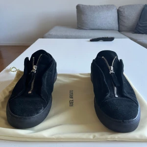 Svarta sneakers Axel Arigato mocka - Svarta sneakers från Axel Arigato i mjuk mocka med cool dragkedja framtill istället för klassisk snörning. Platt sula och rund tå, med detaljer i svart skinn på hälen. Snygg minimalistisk design som passar till allt. Storlek 43 men passar även 42-44 då det är dragkedja 