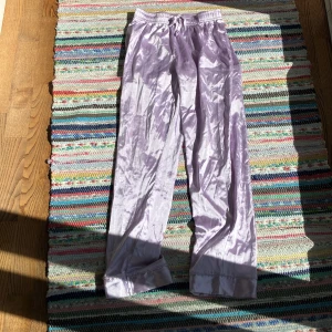 Ljuslila satinglansiga byxor 146/152 - Snygga ljuslila byxor i glansigt satintyg med resår och snörning i midjan. Byxorna har raka ben och breda muddar nertill för en chill vibe. Perfekta för dig som gillar mjuka och bekväma plagg med lite extra shine.