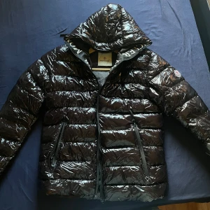 Svart glansig dunjacka från Moncler - Säljer en svart glansig dunjacka från Moncler med huva och dragkedja. Jackan har Moncler-logga på ärmen, två fickor med dragkedja och quiltad design. Perfekt för dig som vill ha en snygg och varm jacka med streetkänsla.