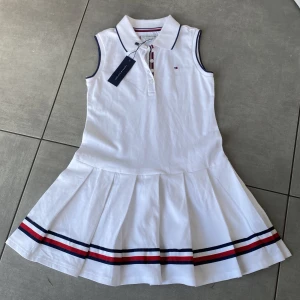 Vit kortklänning Tommy Hilfiger - Supersnygg vit kortklänning från Tommy Hilfiger med klassisk pikékrage och knappar framtill. Ärmlös modell med marinblå och röda ränder längs kjolens kant och vid knappslån. Kjolen är plisserad och klänningen har diskreta Hilfiger-detaljer. Perfekt för en sportig och fräsch look.