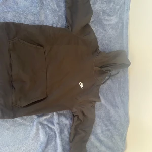 Svart Nike hoodie med huva - Svart hoodie från Nike med klassisk vit logga på bröstet. Tröjan har huva med snörning och en stor känguruficka framtill. Perfekt för chill dagar eller när du vill ha en enkel och stilren look.