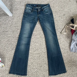 Blå bootcut jeans från Rebelseven - Snygga blå bootcut jeans från Rebelseven med slitningar och ljusare partier framtill. Klassisk femficksmodell med nitar och dekorativa sömmar på bakfickorna. Jeansen har låg midja och är tillverkade i jeansmaterial med stretch. Innerbenslängden är 79 cm❤️ midjemått rakt över är 37 cm
