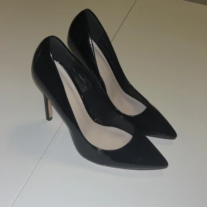 Svarta klackar-Faith - Skitsnygga svarta klackar från Faith i storlek 37 med bred passform. Skorna har en spetsig tå och hög klack, tillverkade i blankt syntetmaterial som ger en riktigt clean look. Perfekta för dig som vill ha en klassisk och stilren vibe. Använd endast 2 gånger.