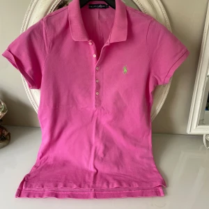 Rosa pikétröja från Ralph Lauren - Klassisk rosa pikétröja från Ralph Lauren med korta ärmar och krage. Tröjan har knappar framtill och den ikoniska gröna polospelaren broderad på bröstet. Perfekt för en sportig och preppy look. Tillverkad i mjuk bomull som andas.den är som som em smal &medium