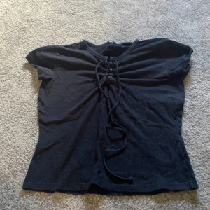 Svart topp med snörning framtill - Svart topp i mjuk bomull med korta ärmar och snygg snörningsdetalj framtill vid halsen. Modellen är enkel men cool, perfekt att styla med jeans eller kjol för en trendig look. Toppens passform är normal och materialet känns bekvämt mot huden.