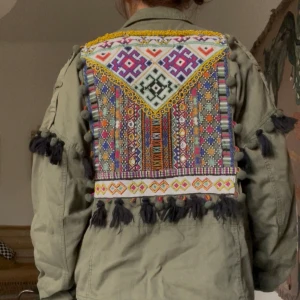Grön jacka med broderad rygg - Unik grön jacka med färgglad broderad ryggpanel och svarta fransar längs ärmar och rygg. Jackan har flera fickor framtill, knäppning med stora knappar och coola detaljer som ger en bohemisk vibe. Perfekt för dig som vill sticka ut med din stil. Den har vissa defekter på baksidan för mer bilder så skriv!!