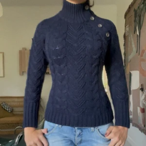 Mörkblå stickad polotröja med knappar - Snygg mörkblå stickad polotröja med kabelstickat mönster och dekorativa knappar på axeln. Tröjan har hög krage, lång ärm och ribbade muddar. Perfekt för dig som gillar en klassisk men trendig look.