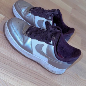 Nike Air Force 1 silver sneakers - Säljer ett par Nike Air Force 1 sneakers i metallic silver med vita detaljer och svart snörning. Skorna har en klassisk rund tå och platt sula, med den ikoniska swoosh-loggan på sidan. Perfekt för dig som vill sticka ut med en clean och modern look.