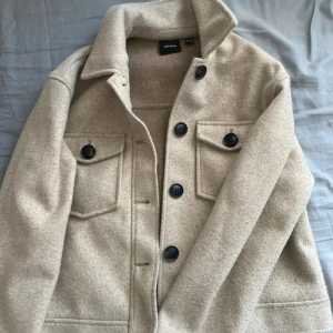 Beige kappa från Vero Moda - Snygg beige kort kappa från Vero Moda med stora svarta knappar och två bröstfickor med lock. Jackan har en rak passform och är gjord i ullblandning, perfekt för lager-på-lager-stil under höst och vår.