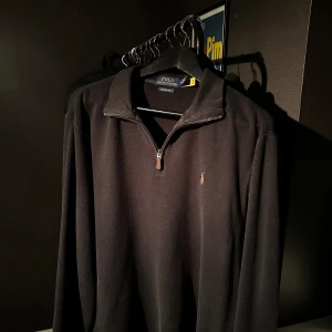 Svart half zip tröja från Polo Ralph Lauren - Svart långärmad tröja från Polo Ralph Lauren med klassisk broderad logga på bröstet. Tröjan har en stilren half zip-dragkedja och ribbad krage. Materialet är mjukt och känns skönt mot huden, perfekt för chill höst- och vinterdagar.