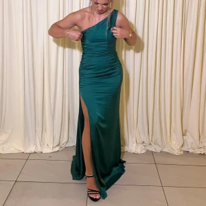 Mörkgrön festklänning klänning med slits - Säljer en elegant mörkgrön långklänning i satin med oneshoulder och hög slits på ena sidan. Klänningen har en figurnära passform och snygga draperingar längs midjan. Perfekt för bal eller fest! Fråga gärna om fler bilder eller mått. Klänningen är endast använd en kväll. Köptes på en klännings butik i Malmö för 2500kr
