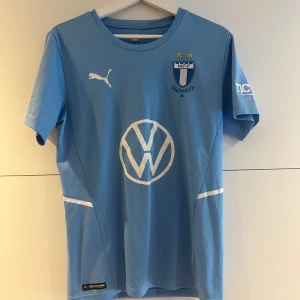 Malmö FF matchtröja Puma ljusblå - Ljusblå Malmö FF fotbollströja från Puma med klubbmärke, två stjärnor och sponsorloggor på bröstet. Kortärmad modell i lätt och ventilerande material, perfekt för träning eller match. Klassisk rund hals och vita detaljer på sidorna.