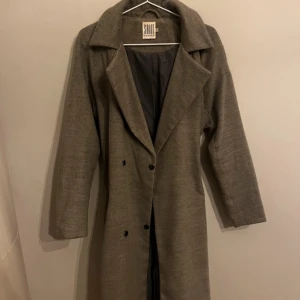 Grå trenchcoat från Saint Tropez - Snygg grå trenchcoat från Saint Tropez med klassisk krage, knappar framtill och bälte i midjan. Jackan har långa ärmar och en stilren look som passar perfekt till lager-på-lager. Fodrad insida för extra komfort och snygg passform.