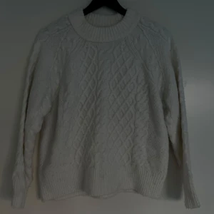 Vit stickad tröja med kabelmönster från Lager157 - Säljer en vit stickad tröja med kabelmönster på framsidan från Lager157. Tröjan har en bred ribbad krage och muddar vid ärmslut och nederkant. Perfekt för dig som gillar mysiga och klassiska vibes under kyliga dagar. 