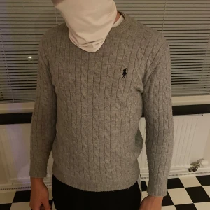 Grå kabelstickad tröja Polo Ralph Lauren - Kabelstickad grå tröja från Polo Ralph Lauren med klassisk rund halsringning och broderad logga på bröstet. Långärmad och i mjuk bomull, perfekt för lager-på-lager. Snygg och tidlös modell som funkar till allt.