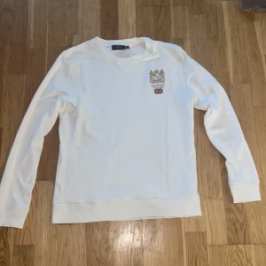Vit sweatshirt från Morris med brodyr storlek S - Vit långärmad sweatshirt från Morris med snyggt broderat emblem och Union Jack-flagga på bröstet. Tröjan har rund hals och ribbade muddar vid ärmslut och nederkant. Perfekt för dig som gillar klassisk stil med en brittisk touch. Tröjan är i mycket bra skick och är i storlek S.