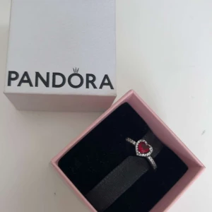 Silverring med röd hjärtformad sten från Pandora - Är du tacksam över en vän eller partner? Säljer då en elegant silverring från Pandora med en röd hjärtformad sten i mitten, omgiven av små glittrande stenar. Ringen levereras i originalask och har en tunn, dekorerad ringskena. Skriv privat för storlek 