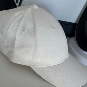 Keps  - Snygg och stilren vit keps i bomull med klassisk design. Kepsen har böjd skärm och justerbart metallspänne baktill för perfekt passform. Passar till många olika outfits och är enkel att matcha.