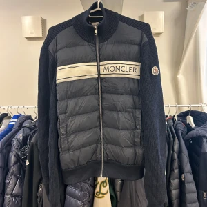 Moncler Logo Tricot Cardigan S - Säljer en svart cardigan jacka från Moncler med stickade ärmar och quiltad front. Jackan har dragkedja, hög krage och Moncler-logga på ärmen. Framsidan pryds av ett vitt band med Moncler-tryck. Perfekt för dig som gillar stilren design. Äkthetsgaranti på samtliga produkter.