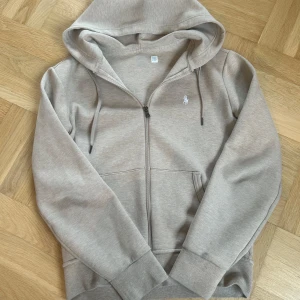 Beige hoodie från Polo Ralph Lauren - Säljer en stilren beige hoodie från Polo Ralph Lauren med dragkedja och huva. Perfekt för en avslappnad look. Mycket bra skick. Den ser grå ut på bilderna men är beige i verkligheten, se på sista bild. Hör av dig vid frågor och funderingar