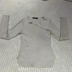 Ljusbeige ribbad topp med cut out från Bik Bok -  ljusbeige ribbad topp från Bik Bok med lång ärm och snygg cut out-detalj vid halsen. Perfekt för dig som vill ha en stilren men ändå unik look. Toppen är figurnära och har rund halsringning.